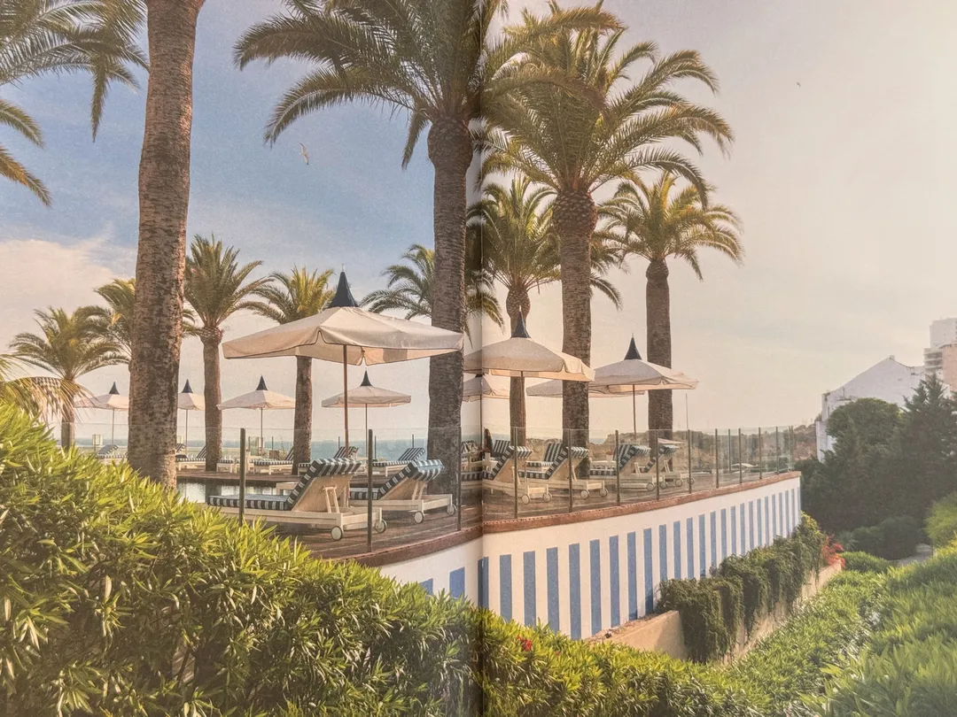 Poolside terrace with palms and white parasols above a blue-and-white striped wall
